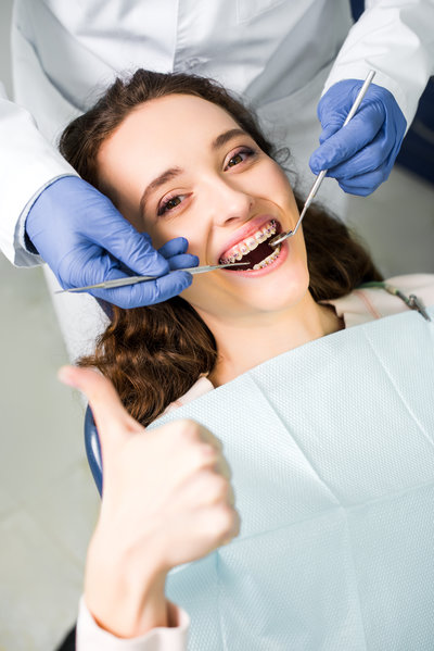 cropped view of dentist in latex gloves examining cheerful woman in braces with opened mouth showing thumb up