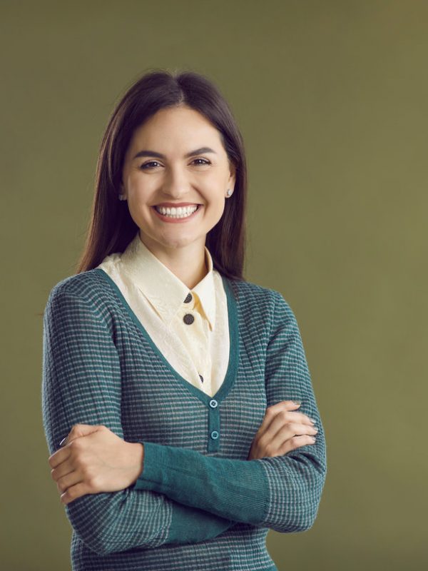 Studio portrait of happy woman with pretty face and positive, charming smile. Cheerful, smiling good-looking young lady in blouse and jumper standing with her arms crossed isolated on green background